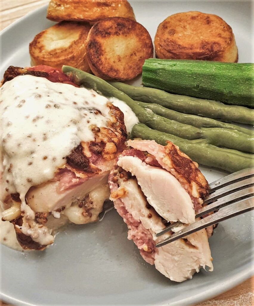 A bacon-wrapped chicken parcel on a plate with vegetables and potatoes.
