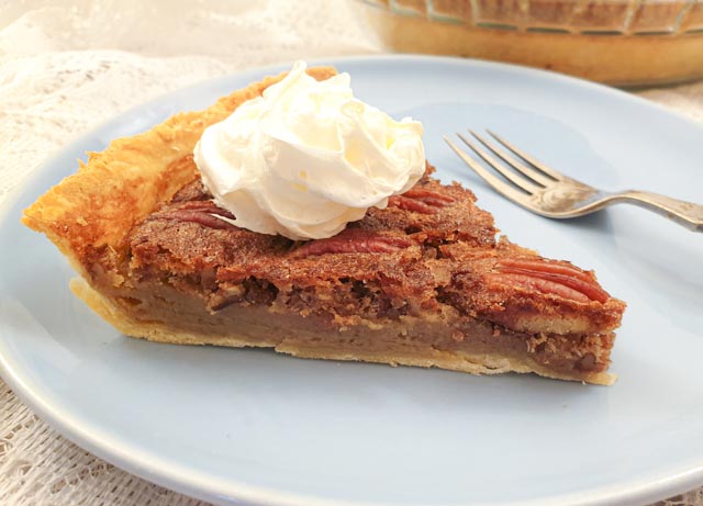 A slice of pecan nut pie, topped with cream, on a plate with a fork.