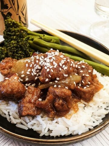 A black dish of sticky asian pork on a pile of white rice, sprinkled with sesame seeds.