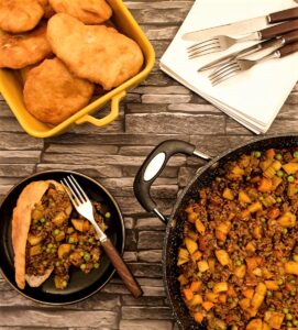 A vetkoek on a plate, filled with curried mince, next to a pan of curried mince and a dish of deep-fried vetkoek.