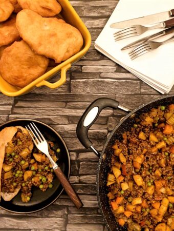 A vetkoek on a plate, filled with curried mince, next to a pan of curried mince and a dish of deep-fried vetkoek.