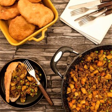 A vetkoek on a plate, filled with curried mince, next to a pan of curried mince and a dish of deep-fried vetkoek.