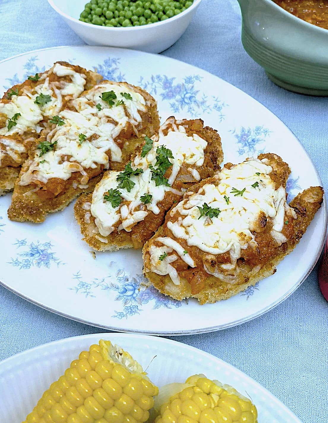 4 pieces of parmesan chicken topped with melted mozzarella on a platter, surrounded by dishes of vegetables.