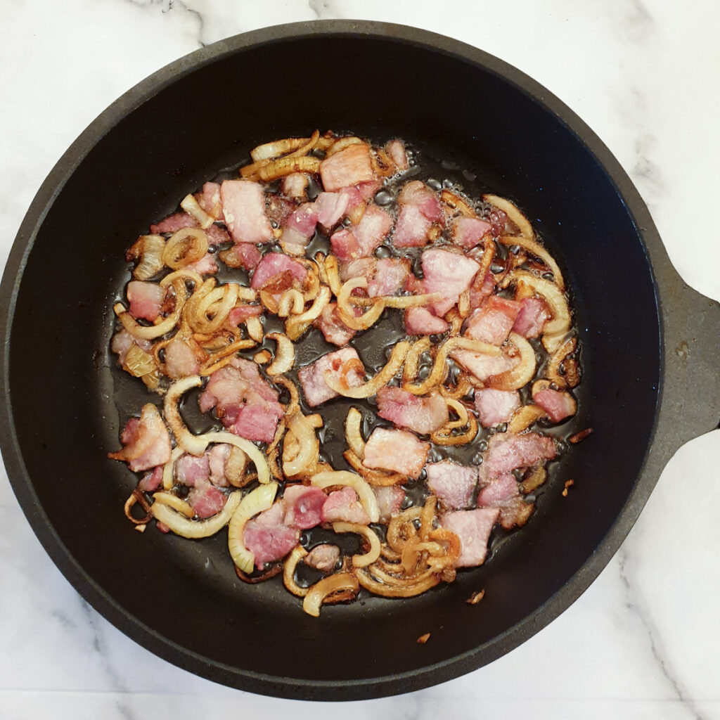 Diced bacon and sliced onions frying in a frying pan.
