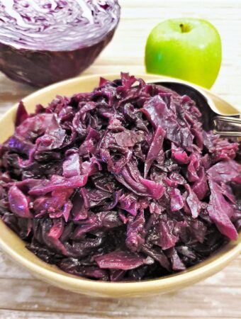 A dish of spicy braised red cabbage with an apple and raw red cabbage in the background.