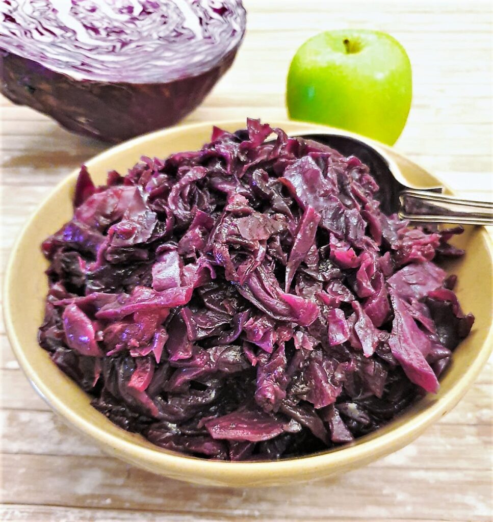 A dish of spicy braised red cabbage with an apple and raw red cabbage in the background.