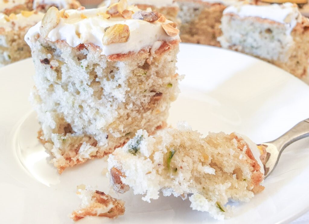 A slice of zucchini and pistachio cake with cream cheese frosting on a plate with a cake fork.