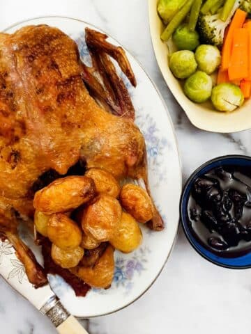 A whole crispy roast duck on a serving plate with roast potatoes, alongside a dish of vegetables and a bowl of cherry sauce.