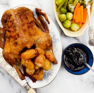 A whole crispy roast duck on a serving plate with roast potatoes, alongside a dish of vegetables and a bowl of cherry sauce.