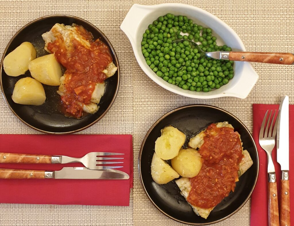Two plates of mediterranean oven-baked cod on the table, with potatoes and a dish of peas.