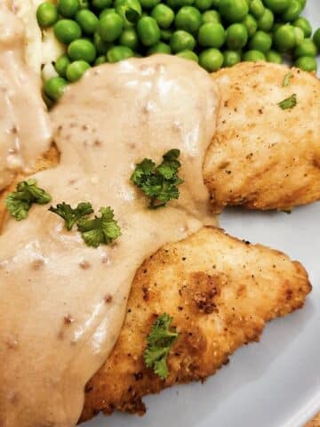 Fried chicken tenderloins on a plate with mashed potatoes and peas, covered with creamy mustard gravy.