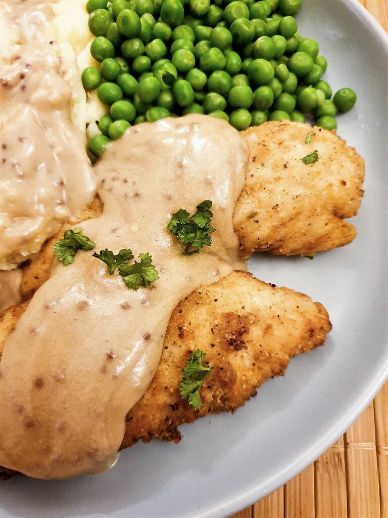 Fried chicken tenderloins on a plate with mashed potatoes and peas, covered with creamy mustard gravy.