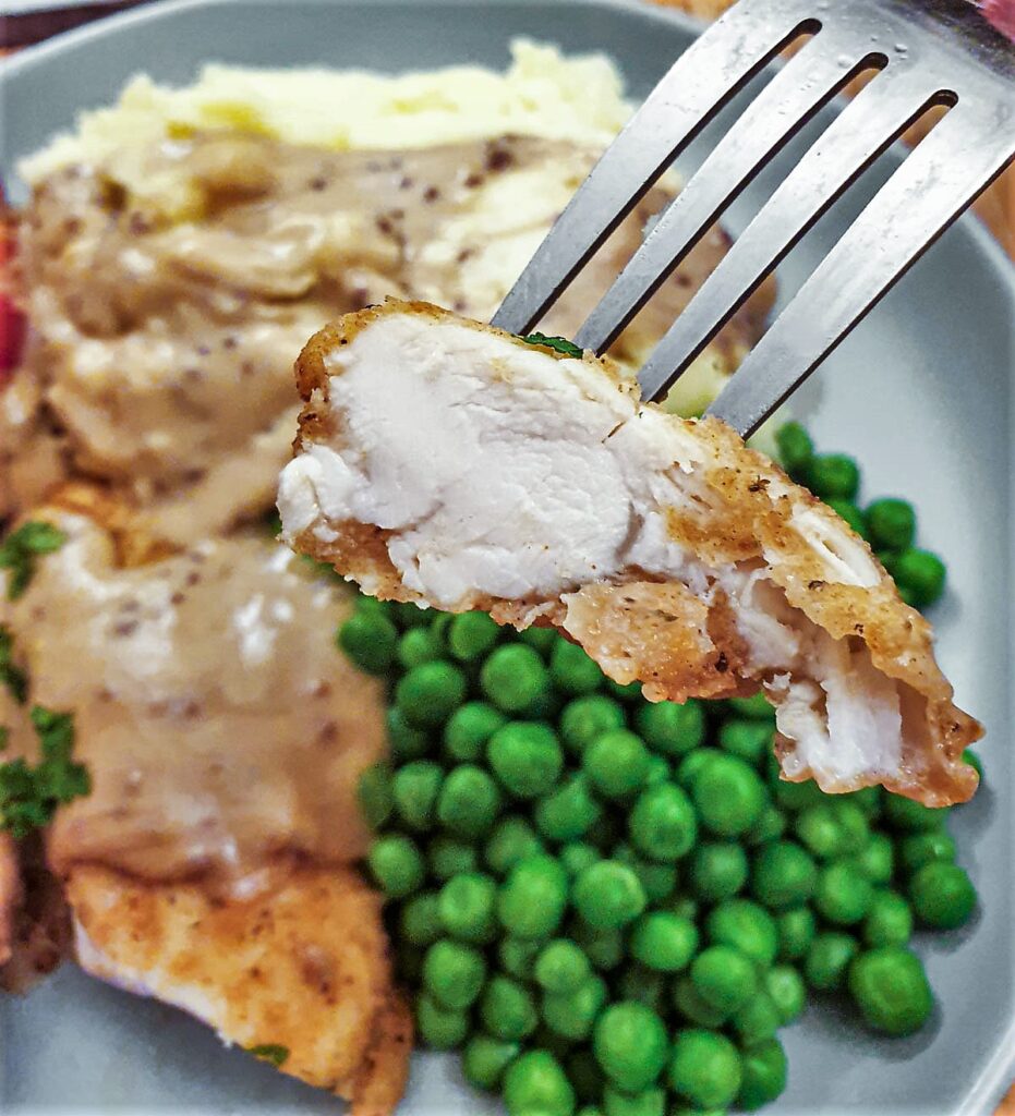 A piece of fried chicken on a fork held above the plate of chicken, mashed potatoes and peas.