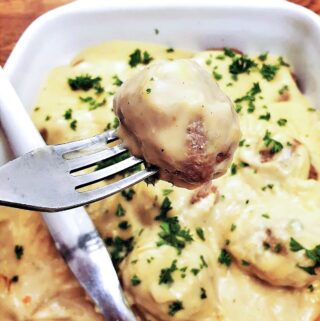 A Swedish meatball on a fork, above a dish of meatballs covered with creamy mustard sauce.