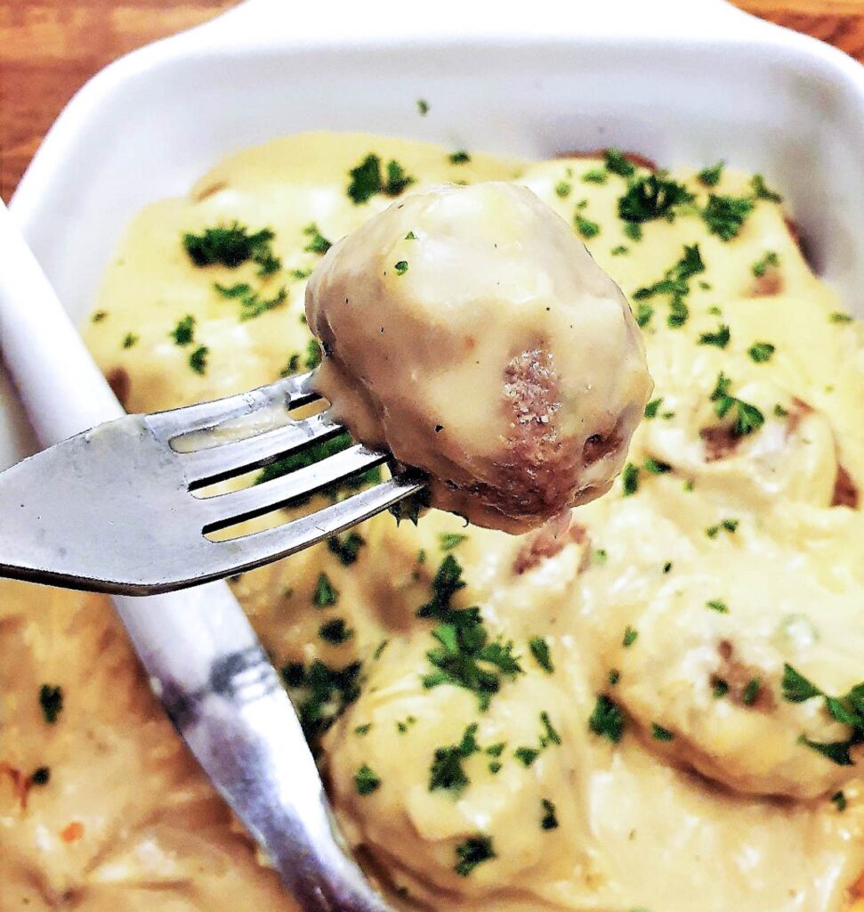 A Swedish meatball on a fork, above a dish of meatballs covered with creamy mustard sauce.