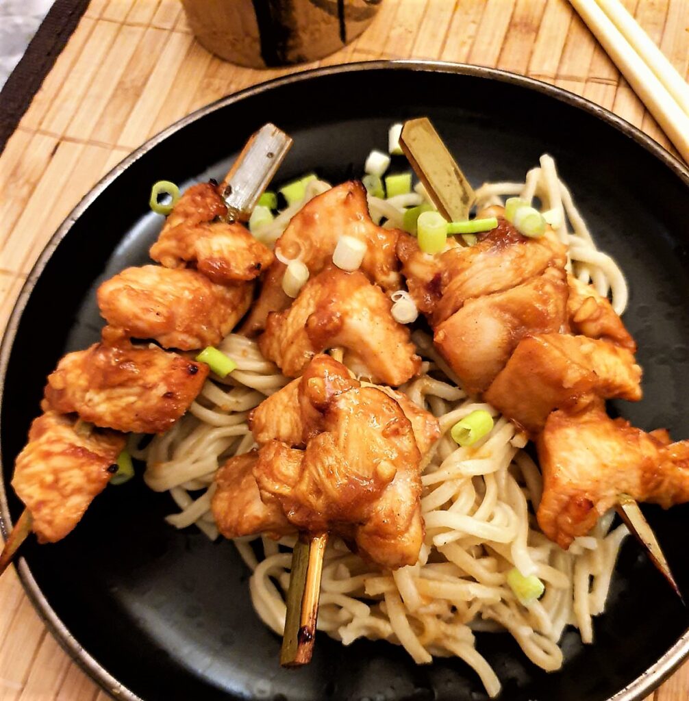 Three sticky chicken skewers arranged on a bed of pasta and garnished with finely chopped spring onions.