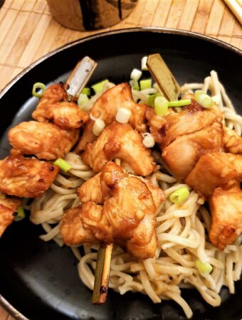 Three sticky chicken skewers arranged on a bed of pasta and garnished with finely chopped spring onions.