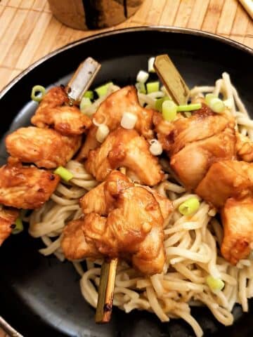 Three sticky chicken skewers arranged on a bed of pasta and garnished with finely chopped spring onions.