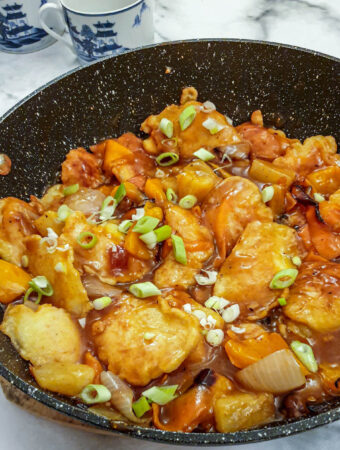 Overhead shot of a pan of sweet and sour chicken pieces garnished with spring onions in a thick sticky sauce.