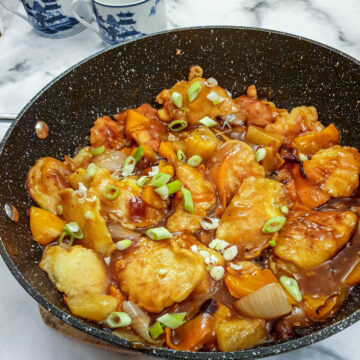Overhead shot of a pan of sweet and sour chicken pieces garnished with spring onions in a thick sticky sauce.