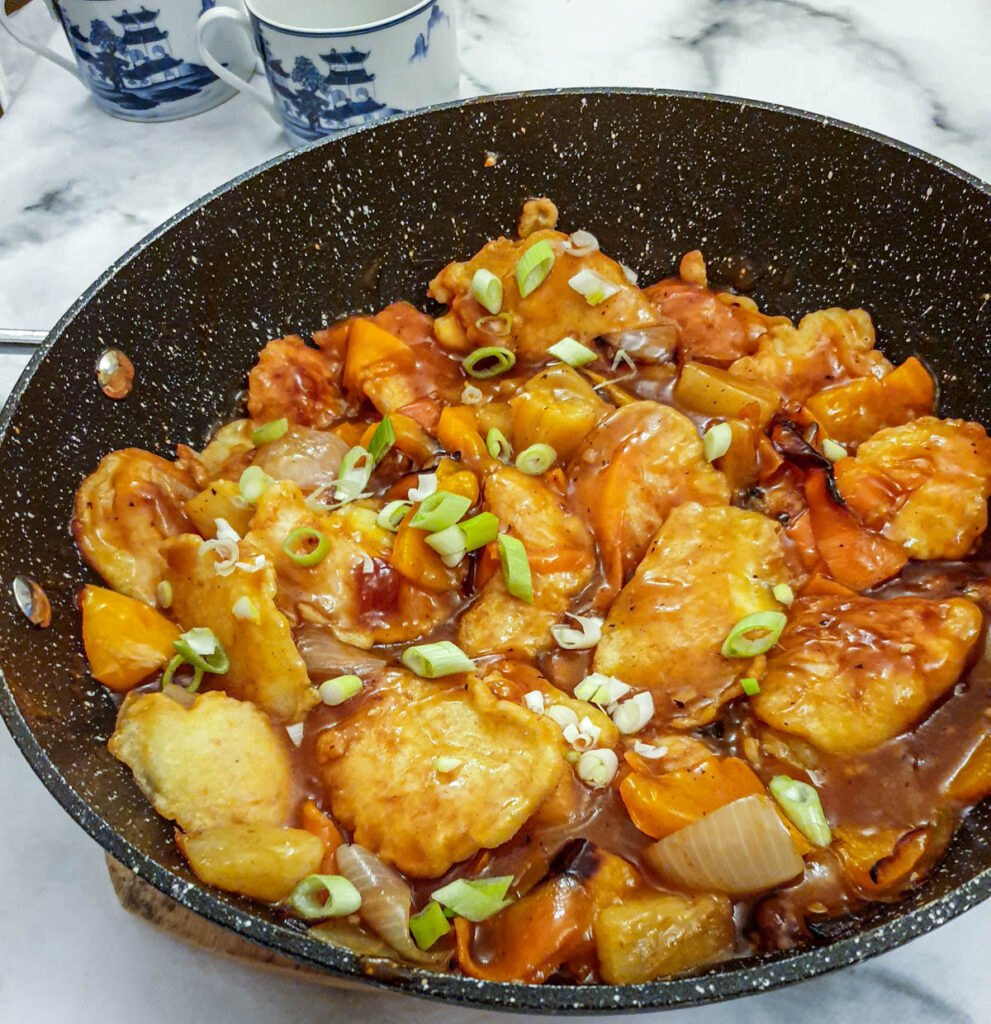 Overhead shot of a pan of sweet and sour chicken pieces garnished with spring onions in a thick sticky sauce.