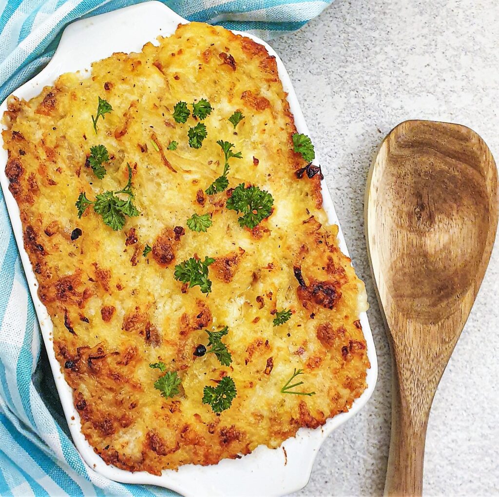 A large casserole dish of golden baked potatoes Romanoff next to a wooden spoon.