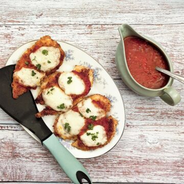 Servings of chicken parmigiana on a plate, next to a dish of marinara sauce.