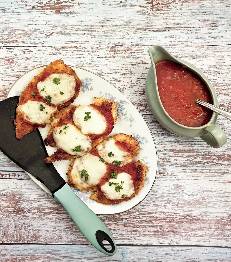 Servings of chicken parmigiana on a plate, next to a dish of marinara sauce.