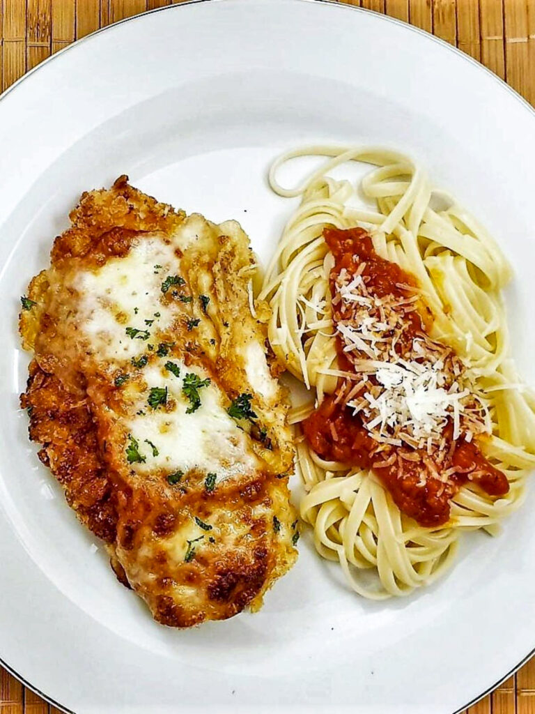 A crispy chicken parmigiana on a plate with a pile of linguine covered with marinara sauce.