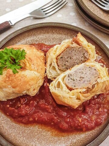 Two meatball wellingtons on a bed of marinara sauce with one cut open to show the inside.