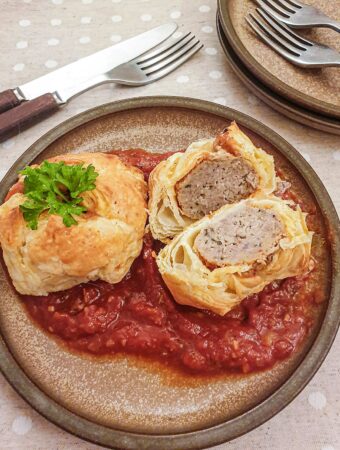 Two meatball wellingtons on a bed of marinara sauce with one cut open to show the inside.