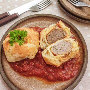 Two meatball wellingtons on a bed of marinara sauce with one cut open to show the inside.