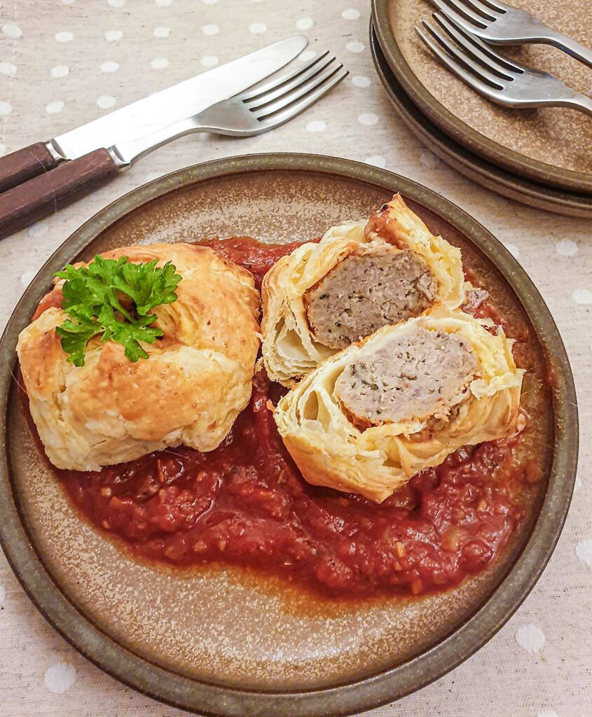 Two meatball wellingtons on a bed of marinara sauce with one cut open to show the inside.