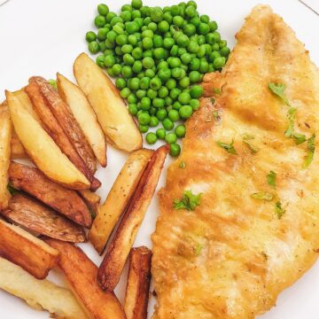 A golden pan-fried fish fillet on a plate with chips and peas.