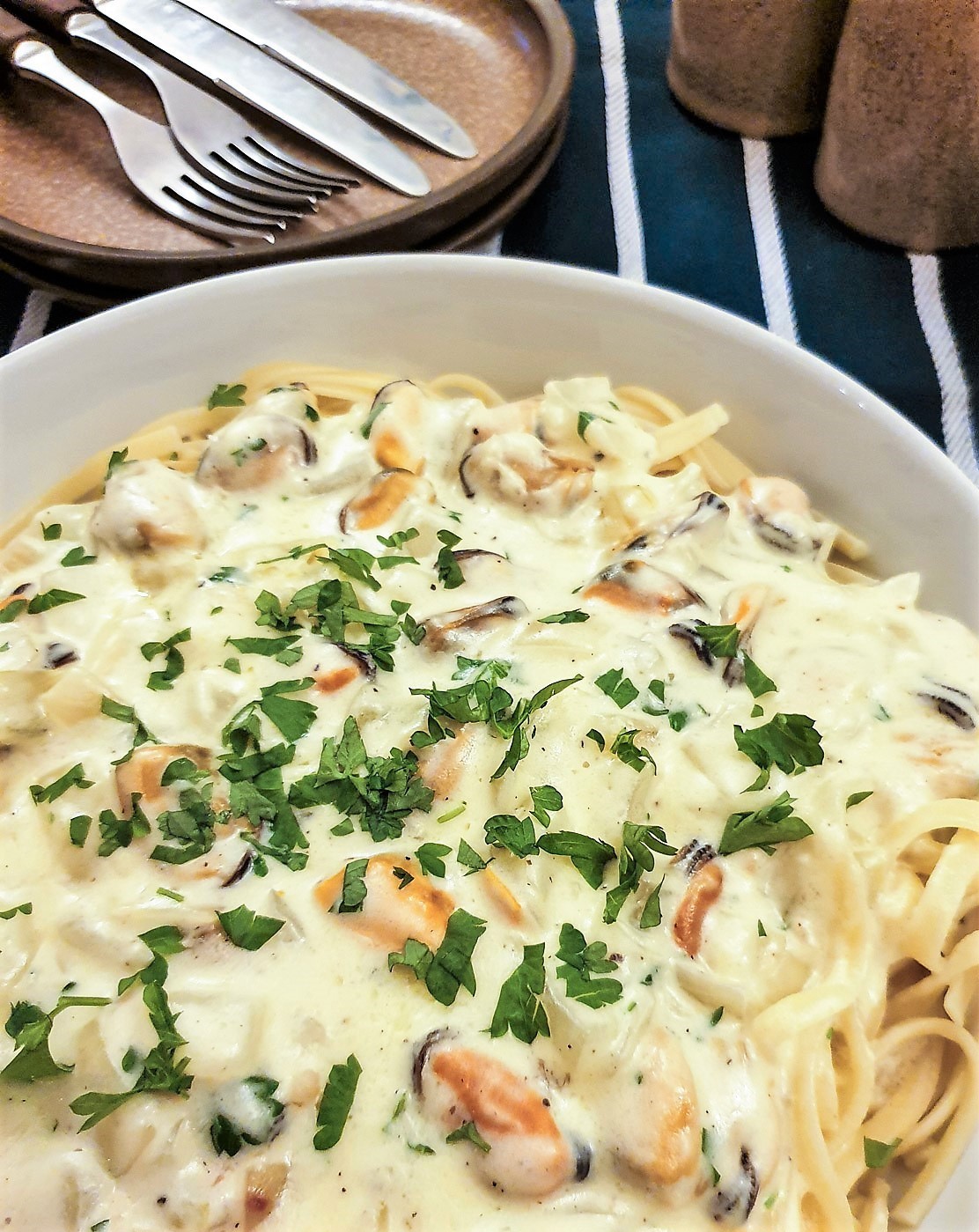 A dish of mussel pasta sprinkled with chopped parsley on a table in front of a plate holding knives and forks.