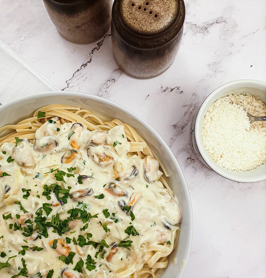 A plate of creamy mussel pasta, sprinkled with chopped parsley, next to  dish of grated parmesan cheese.