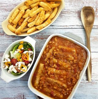 A dish of sausage and bean casserole on a table alongside dish of crispy french fries and a green salad.