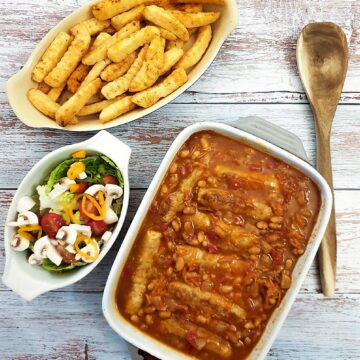 A dish of sausage and bean casserole on a table alongside dish of crispy french fries and a green salad.