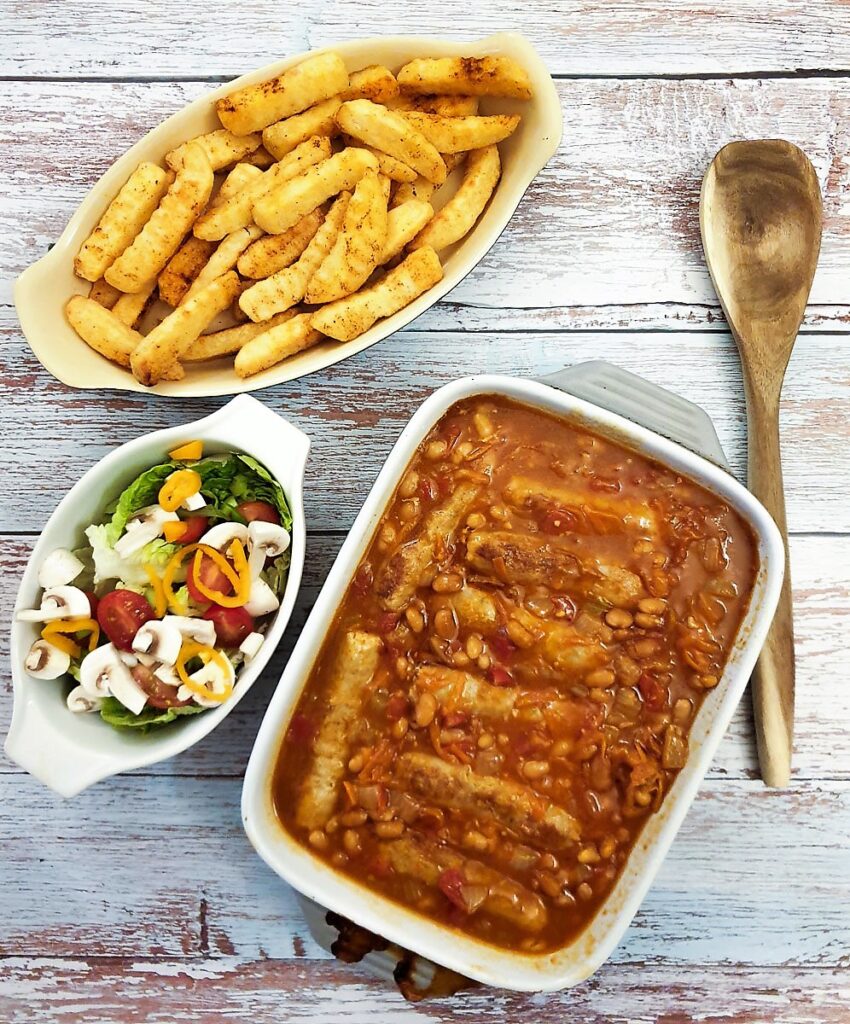 A dish of sausage and bean casserole on a table alongside  dish of crispy french fries and a green salad.