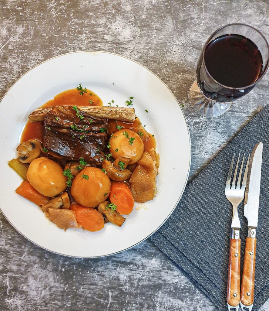 A plate of beef short rib and vegetables next to a glass of red wine