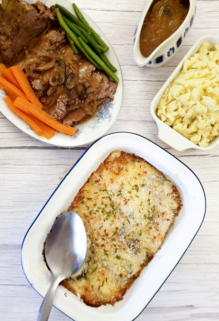 A dish of cauliflower and broccoli cheese sauce with a plate of roast meat and a side of mashed potatoes and gravy.