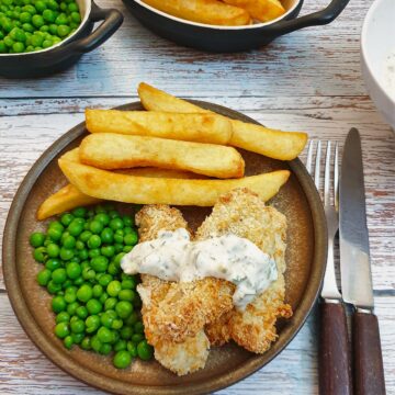 Fish fingers covered in tartare sauce on a plate with french fries and peas.