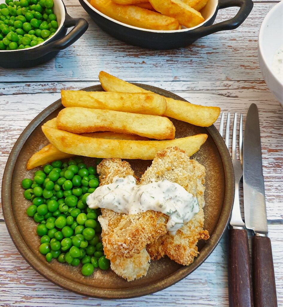 Fish fingers covered in tartare sauce on a plate with french fries and peas.