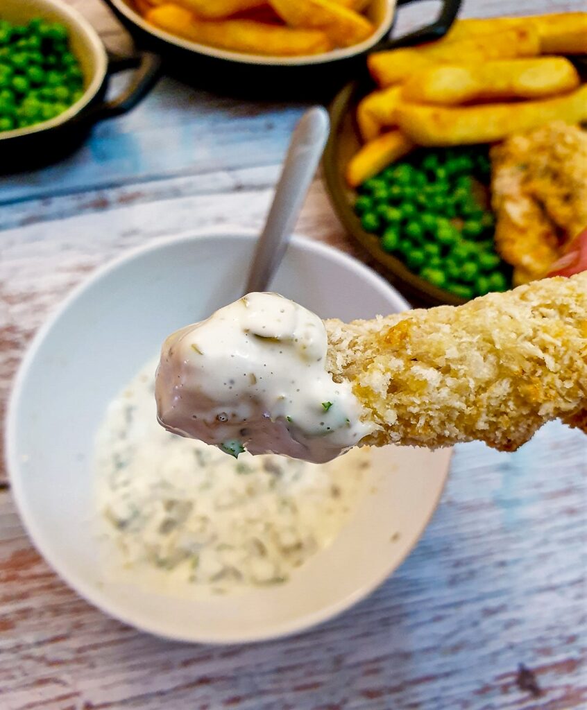 A crispy fish finger being dipped in a bowl of tartare sauce.