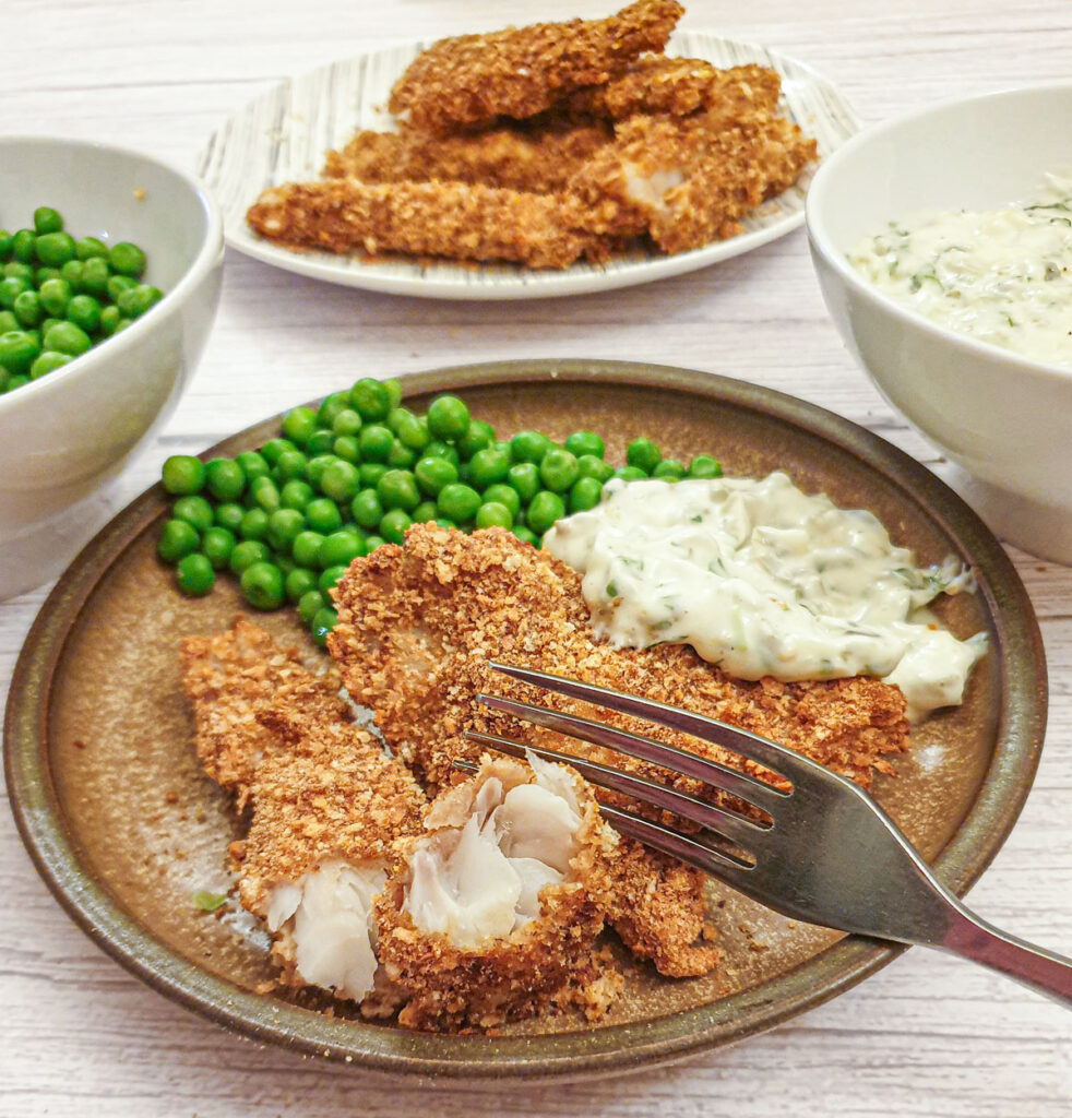 Fish fingers that have been coated in toasted breadcrumbs on a plate with peas and tartare sauce.