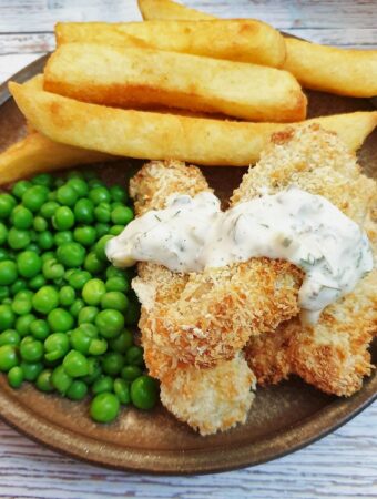 Fish fingers covered in tartare sauce on a plate with french fries and peas.