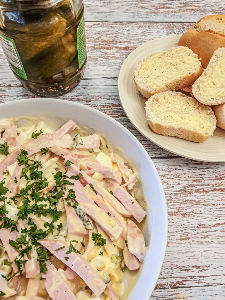 A dish of fleischsalat next to a jar of pickled gherkins.