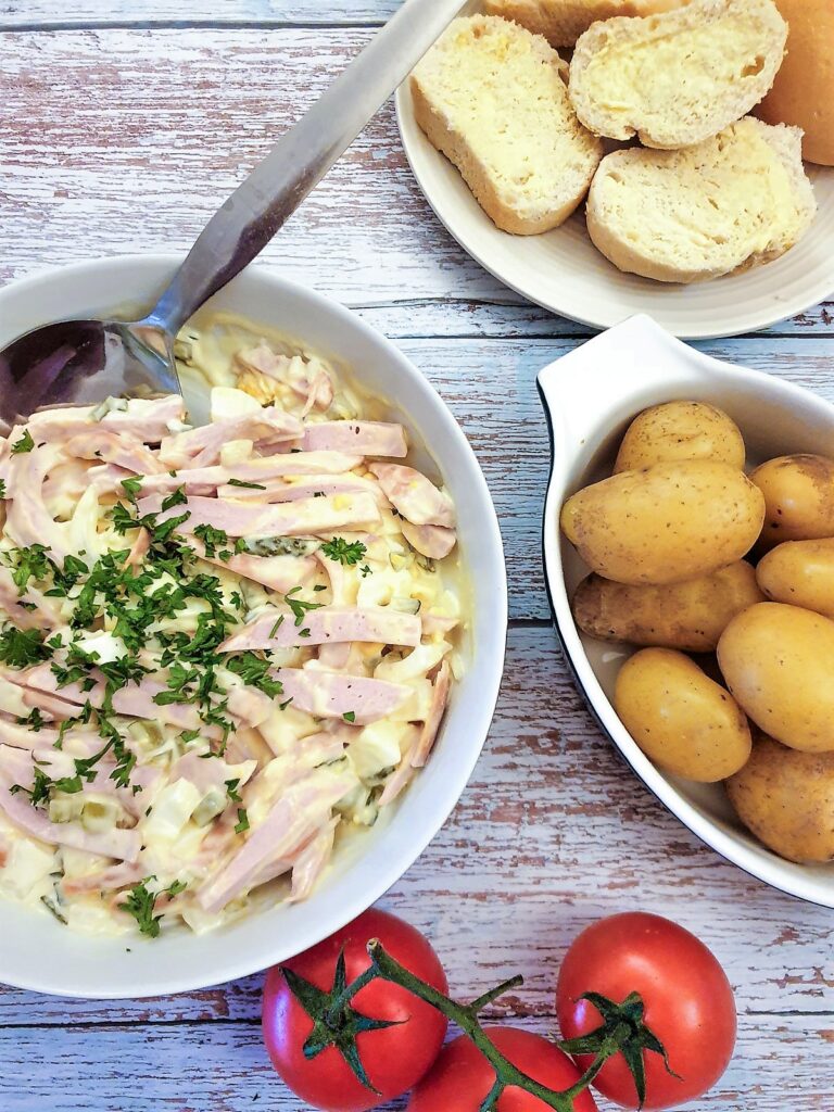 A dish of German meat salad next to a bowl of potatoes and some crusty bread.