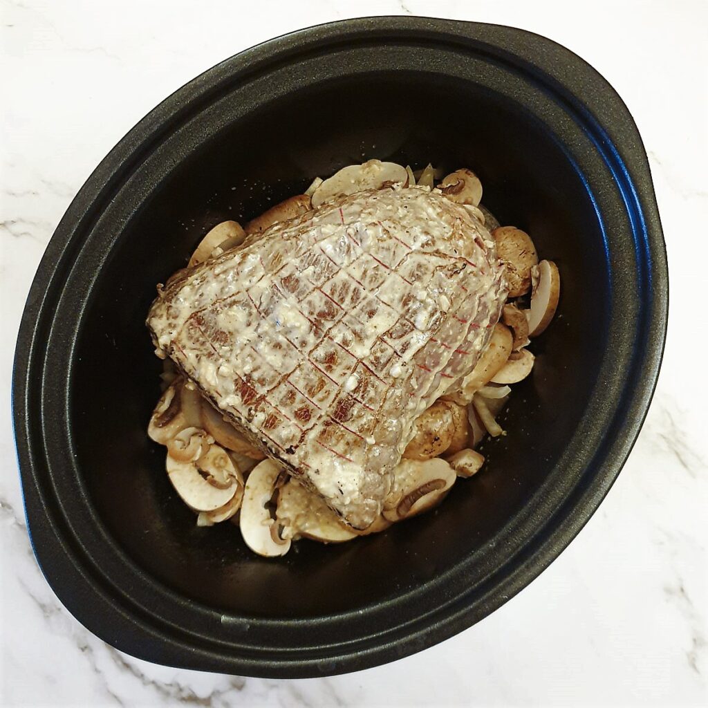 Topside of beef on a bed of mushrooms and onions in the slow-cooker ready to be cooked.