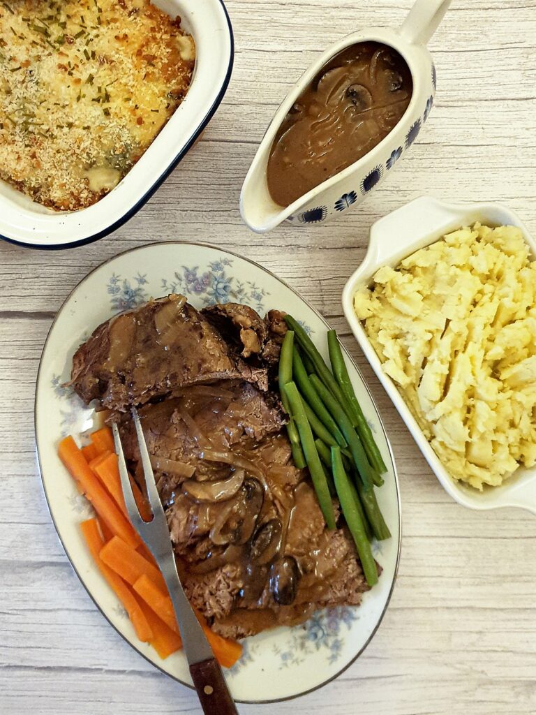 A serving plate of sliced beef topside, with vegetables and a jug of gravy.